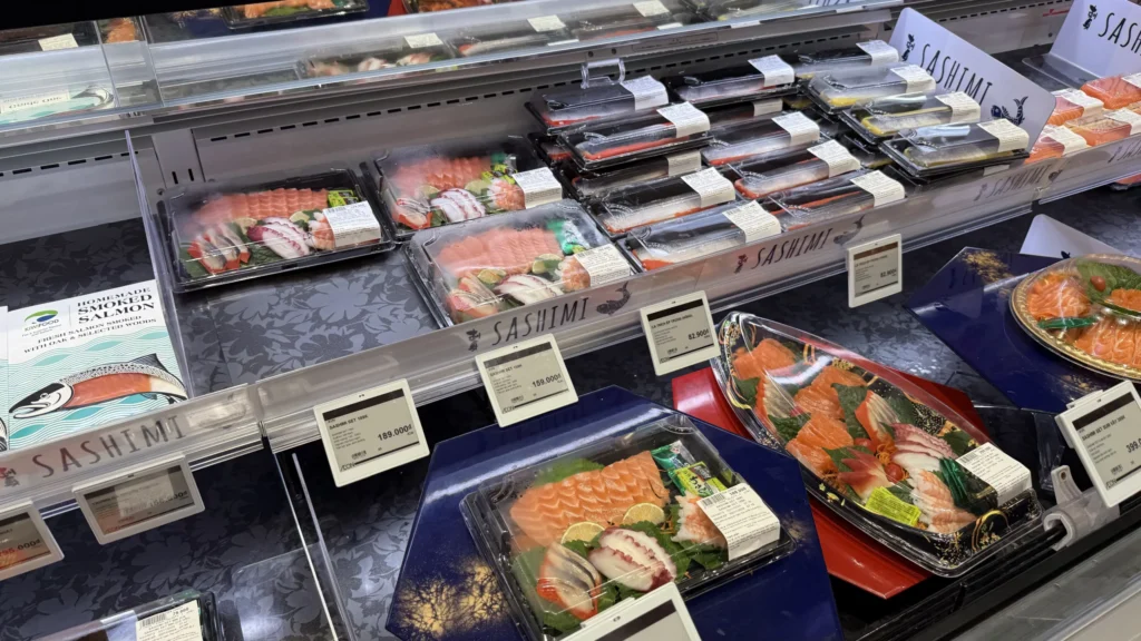 イオンモール フエの食品売り場の刺身コーナー。サーモンやタコの刺身セットが並び、電子値札に159,000ドンなどの価格が表示されている